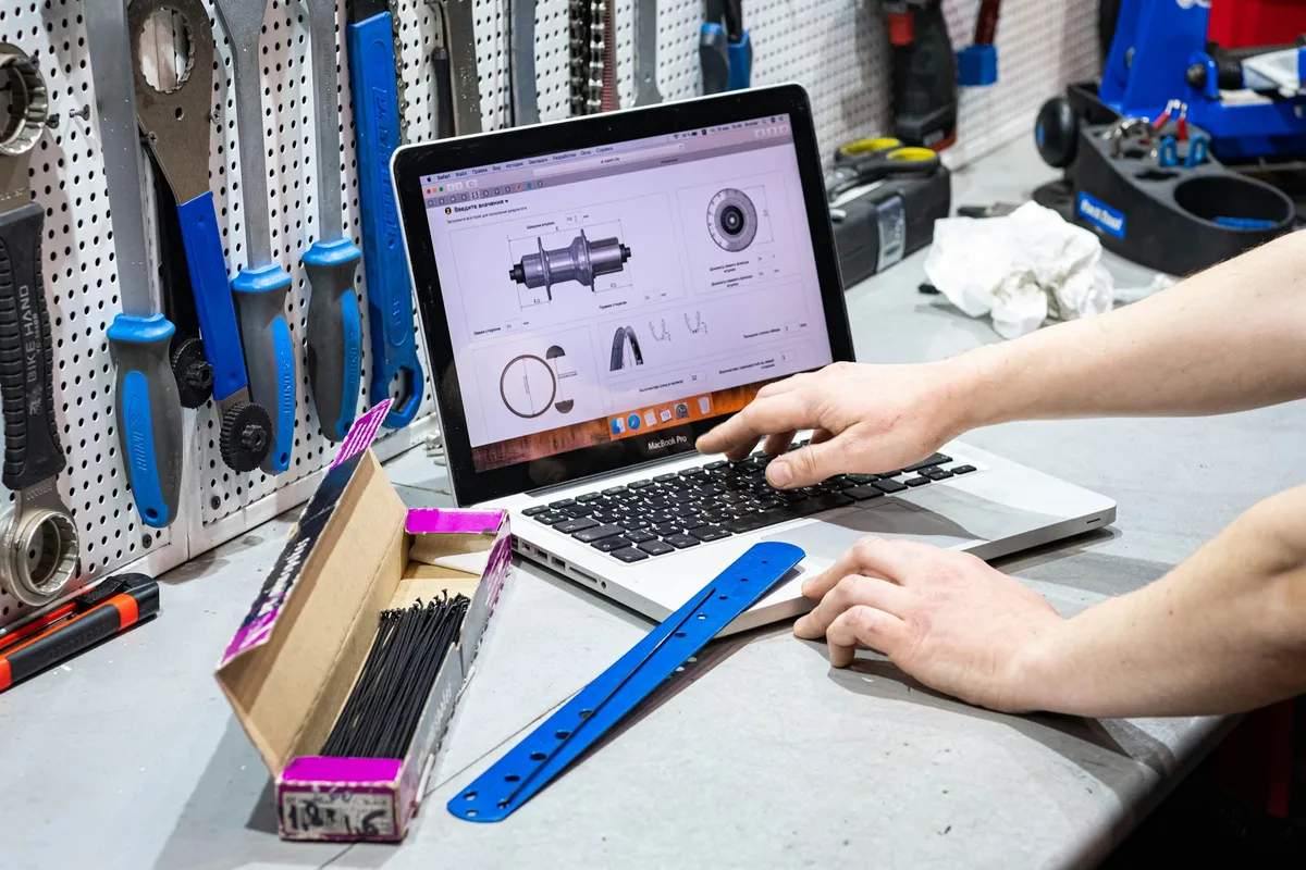 Hands at a laptop on a workbench in a bike workshop, tools on the wall behind