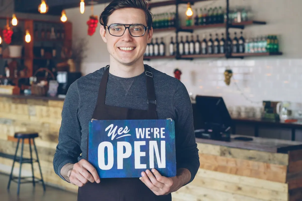 A smiling shopkeeper in an apron holding up a Yes, we're open sign