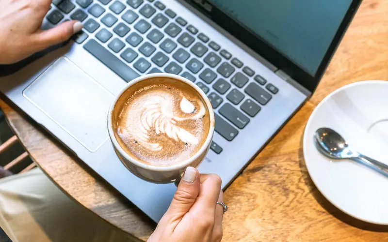 Hands holding a coffee beside an open laptop on a wooden table