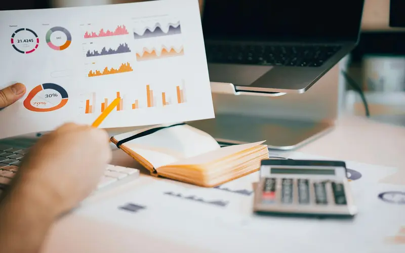 A hand holding printed charts over a laptop and calculator