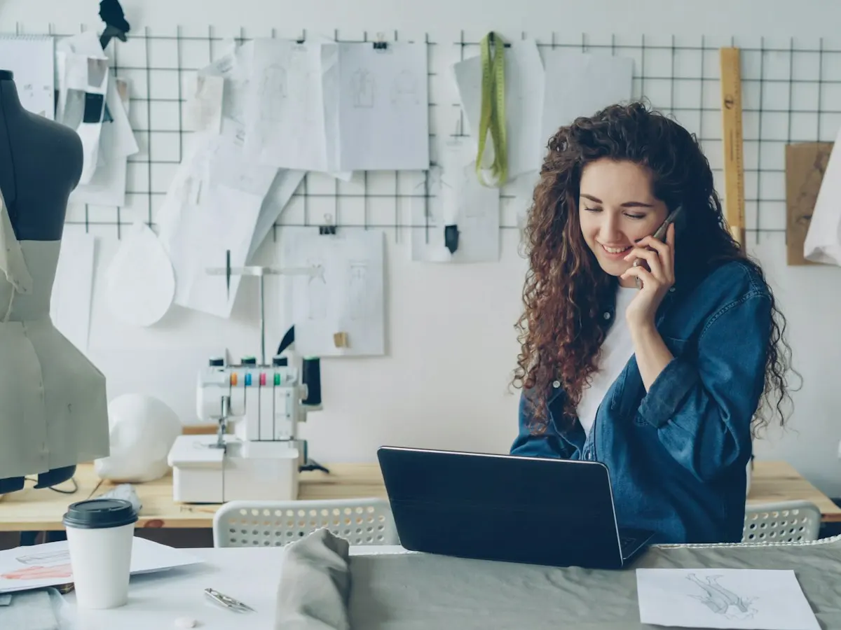 A designer on the phone at her studio workbench, laptop open