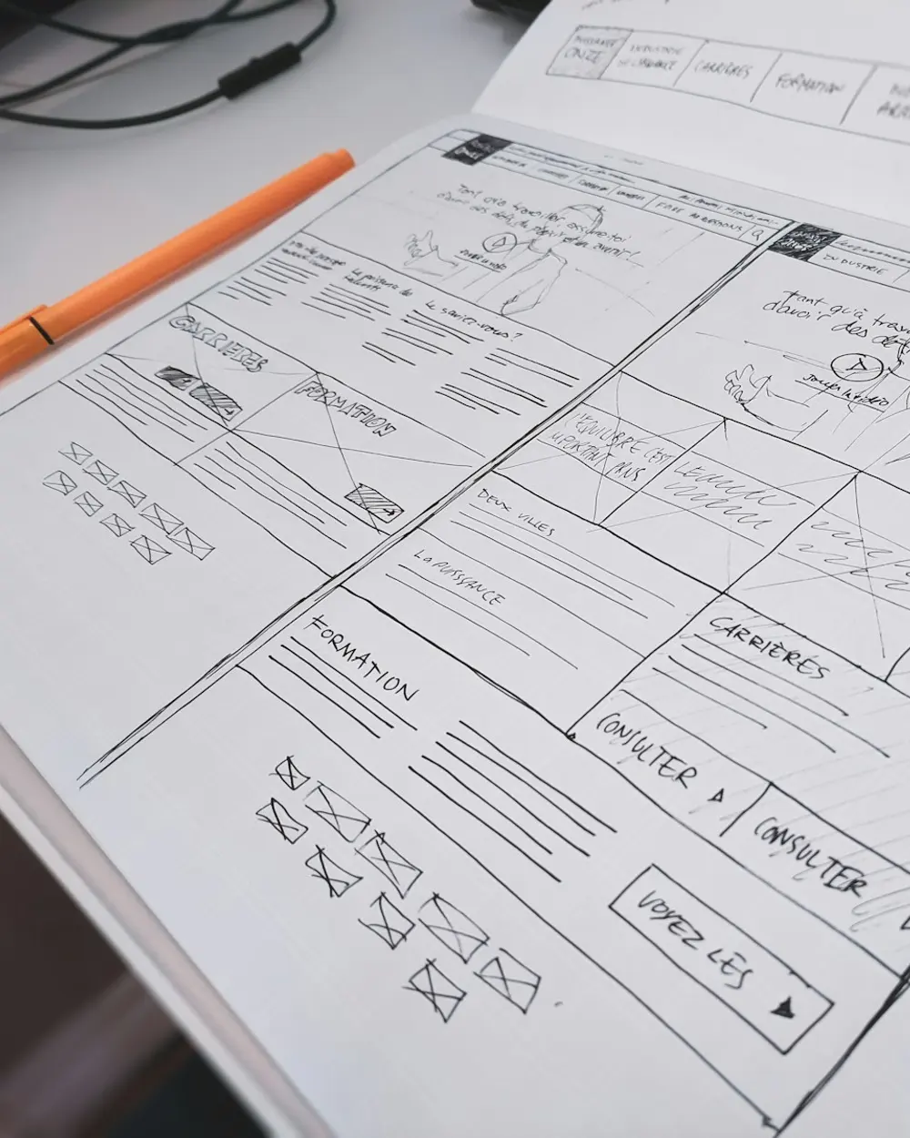 A hand-drawn page layout for a website, sketched on paper beside an orange pen